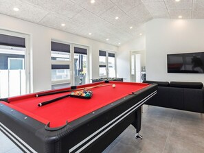 A pool table with balls, a sofa, and a television in a room with windows and a ceiling with recessed lighting.