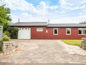 A red single-story house with a white garage door, a brick driveway, and a stone wall with the number 7.