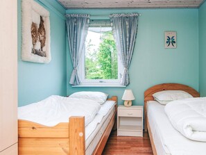 A bedroom with two wooden beds, a window with curtains, a bedside table with a lamp, and a wall-mounted artwork.