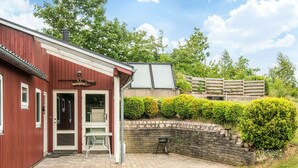 A red building with a door and windows, a small outdoor table, and a stone wall with greenery. - 6 Person Holiday Home in Ebeltoft (Ebeltoft)