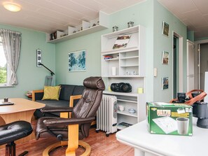 Un espace de vie compact comprenant un canapé, un fauteuil, une table et un radiateur. On peut voir des étagères avec divers objets et une boîte marquée du nom « HATZY ».