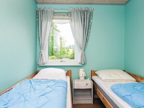 A room with two beds, a window with curtains, a bedside table with a lamp, and a view of greenery outside. - 6 Person Holiday Home in Ebeltoft (Ebeltoft)