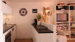 Ett kompakt kök med vita skåp, en svart bänkskiva och en inbyggd ugn. Det finns en rund väggklocka, en liten krukväxt och en bokhylla med diverse föremål.