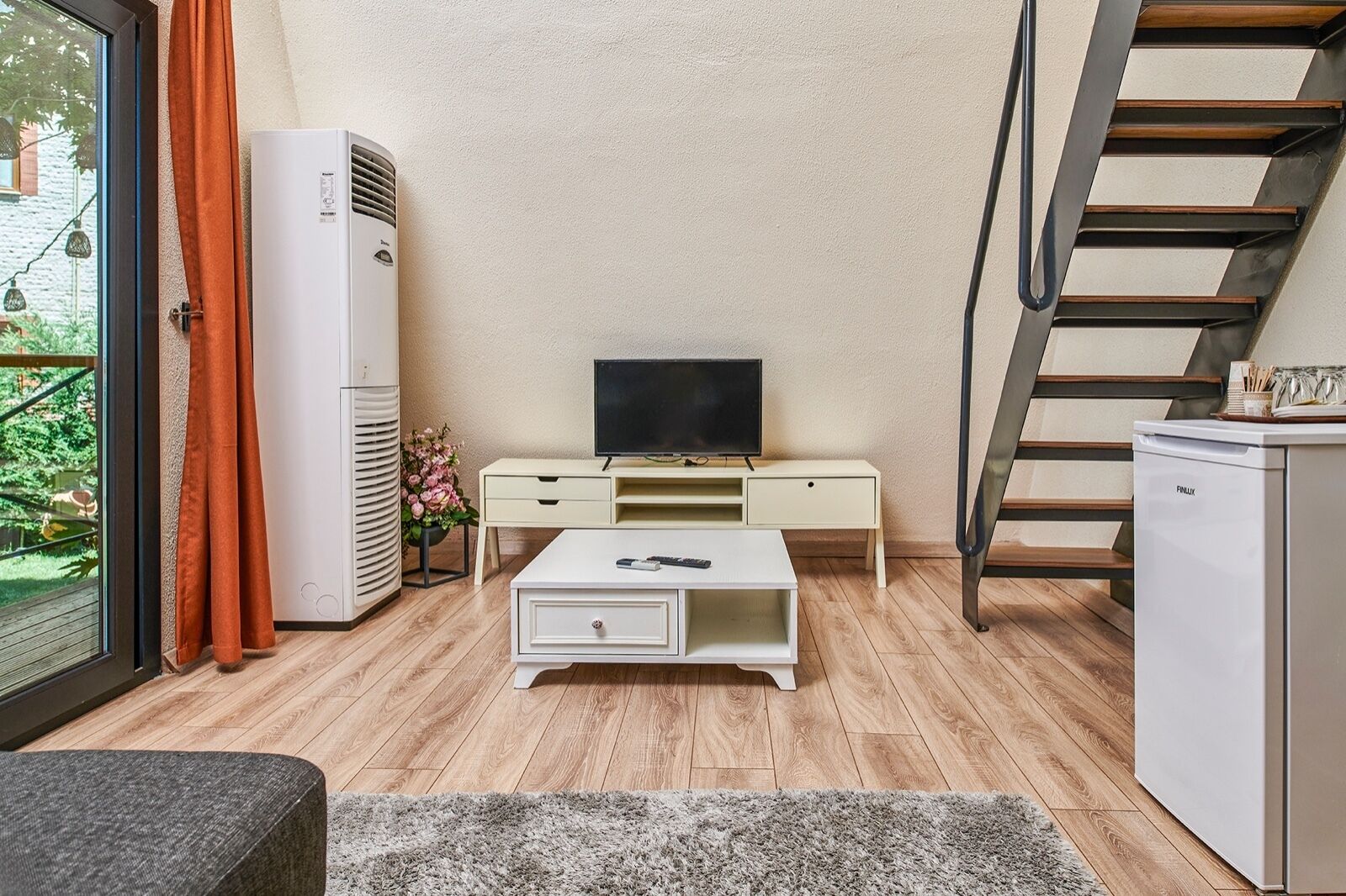 Un espacio habitable reducido con una televisión sobre una mesita blanca, un refrigerador pequeño y una escalera.