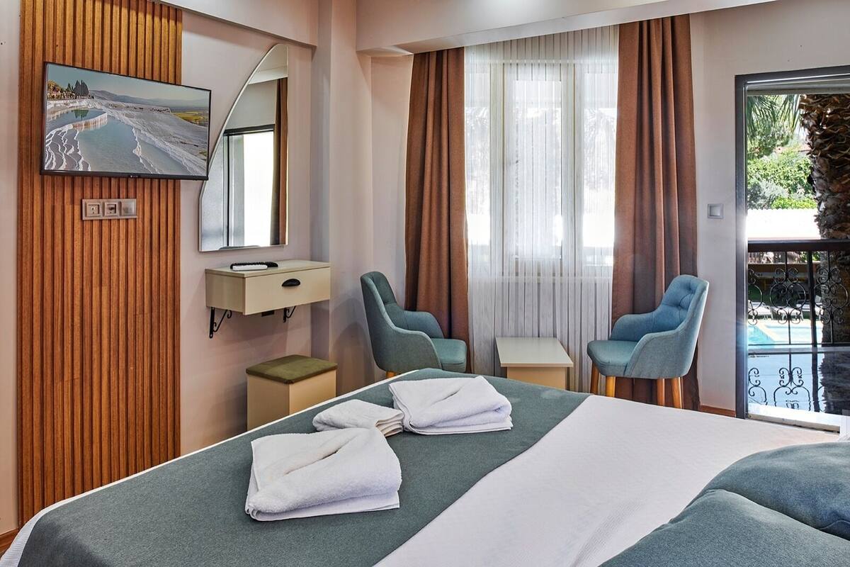 Habitación de hotel con una cama, dos sillas, una mesita, televisor montado en un panel de madera, un espejo y una ventana con cortinas.