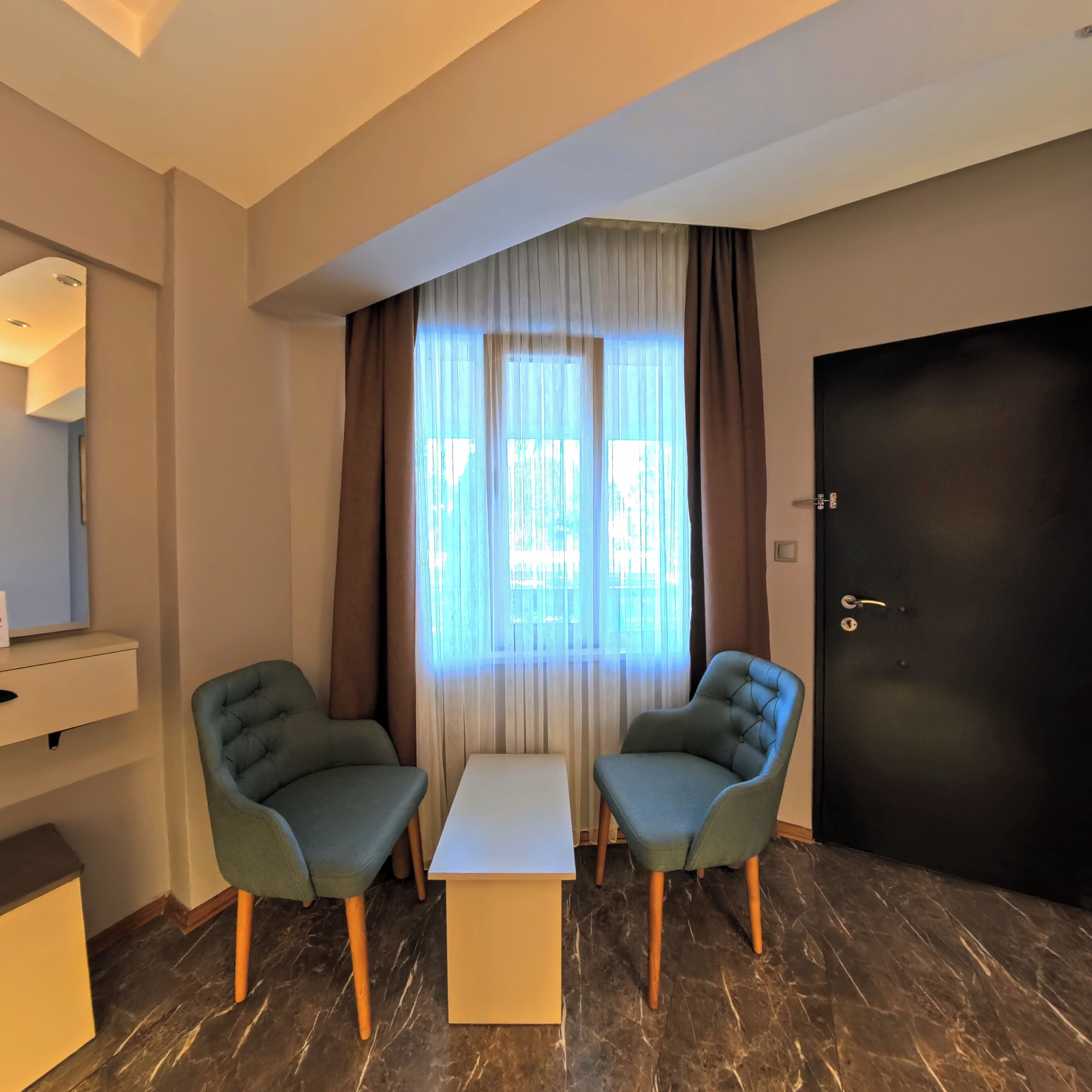 A hotel room with two armchairs, a small table, a dark door, and a window with sheer curtains.