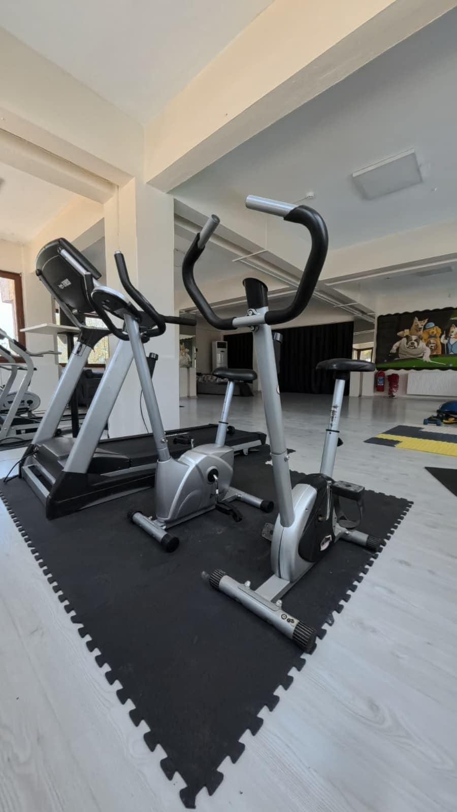 Un gimnasio con tres bicicletas estáticas, una colchoneta negra y un suelo blanco.