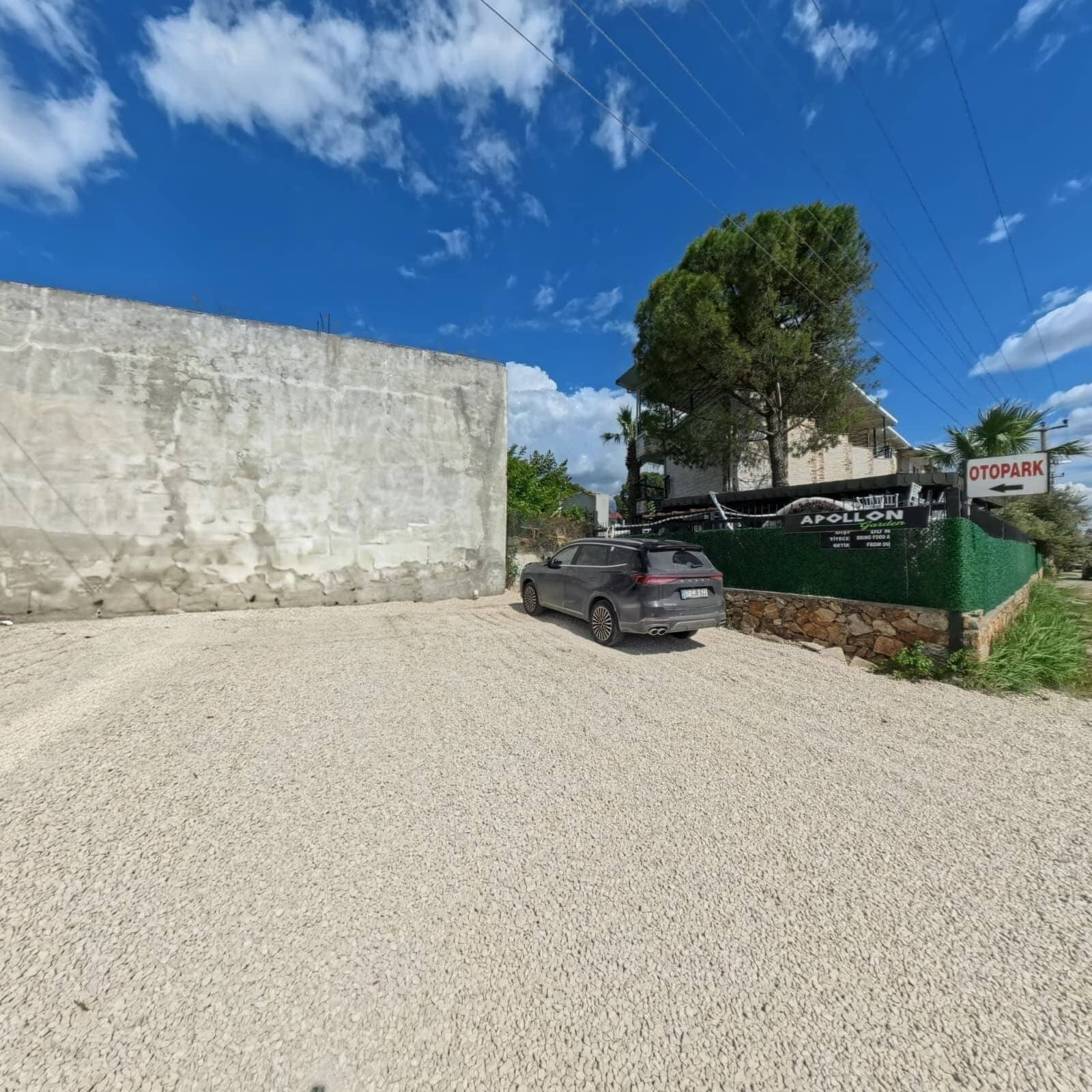 A gravel driveway, a parked SUV, a concrete wall, and a sign for 'A POLLON Garden' with an arrow pointing left to 'OTOPARK'.