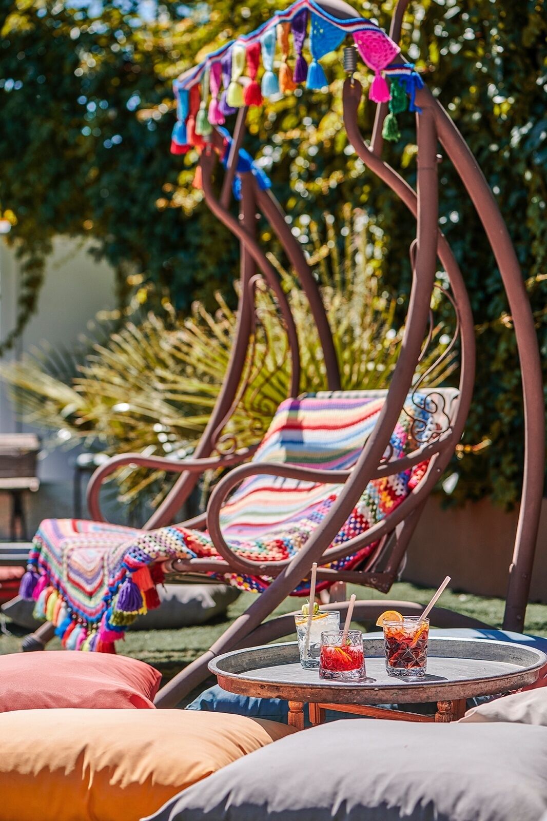 A metal chair with colorful woven seat and backrest, a round table with drinks, and a garden background.