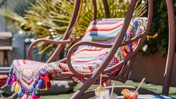 A metal chair with colorful woven seat and backrest, a round table with drinks, and a garden background.