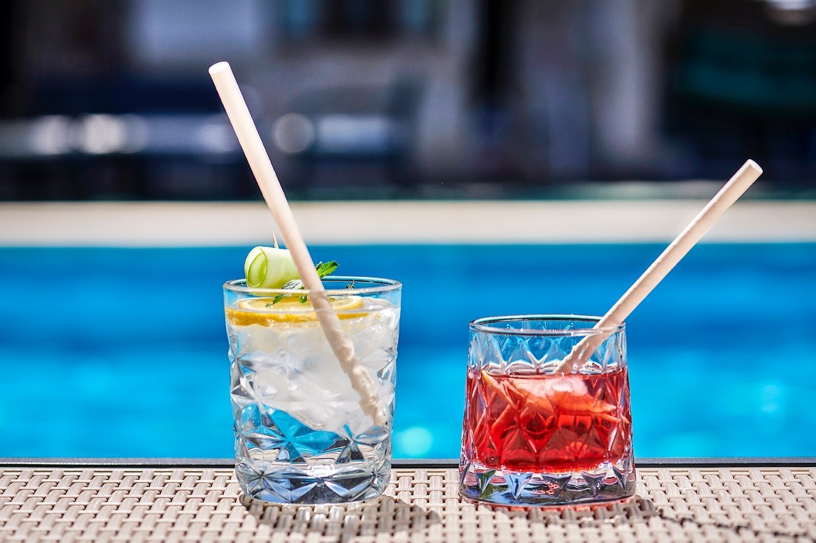 Two glasses of iced drinks with straws, one with a lemon slice garnish, placed on a woven surface by a pool.
