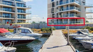 A modern multi-story building with balconies, a dock with boats, and a clear sky.