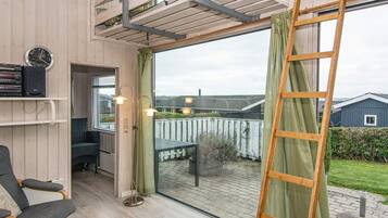 A room with a wooden floor, a chair, a table, a ladder, and a window with a view of a house and garden.