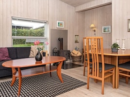 Una sala moderna con un sofá, una mesa de comedor de madera y sillas. Hay una chimenea, un ventanal con vistas a follaje y una alfombra estampada.