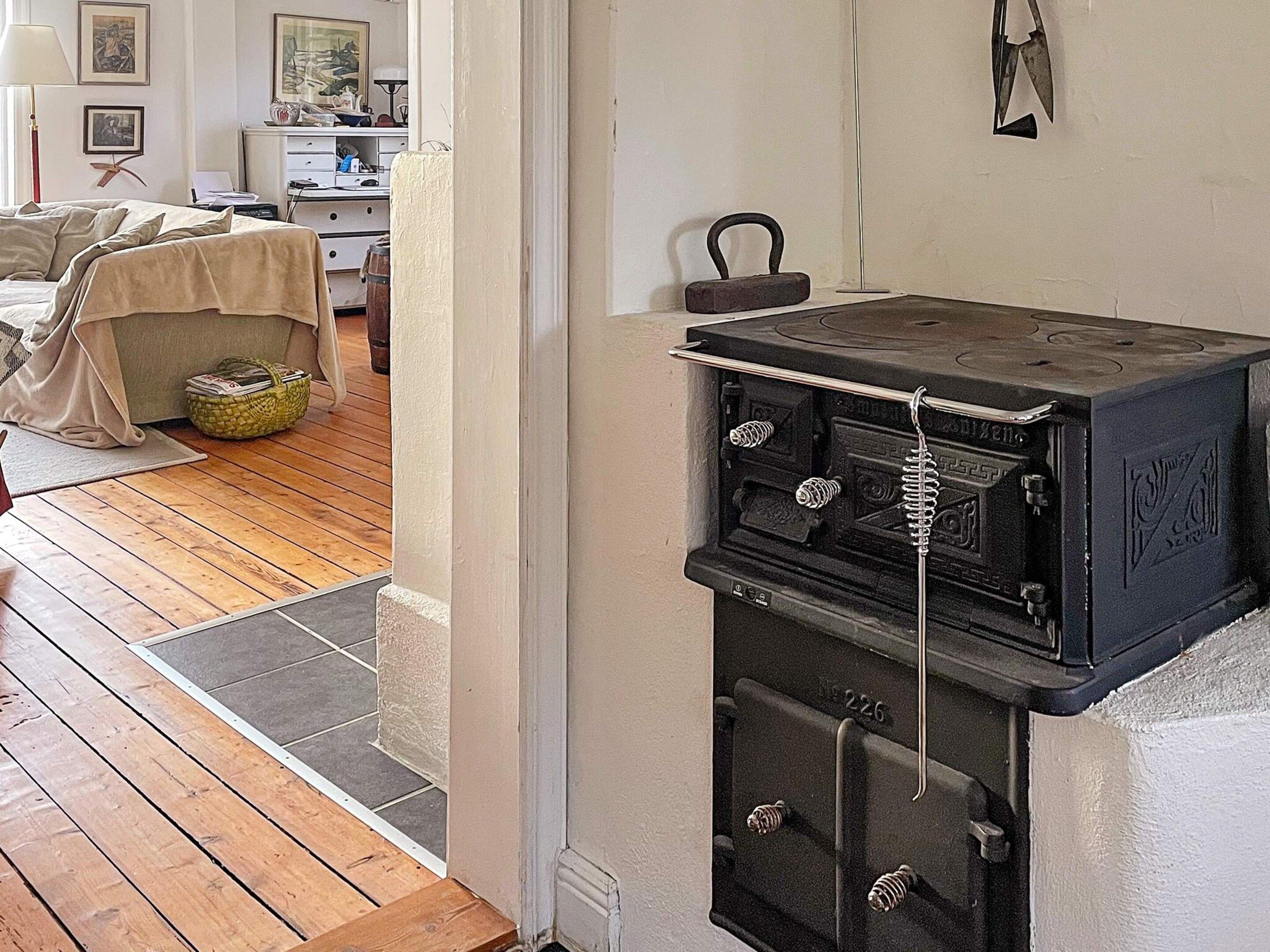 A room with a wooden floor, a bed with a beige cover, a small table, and a vintage stove.