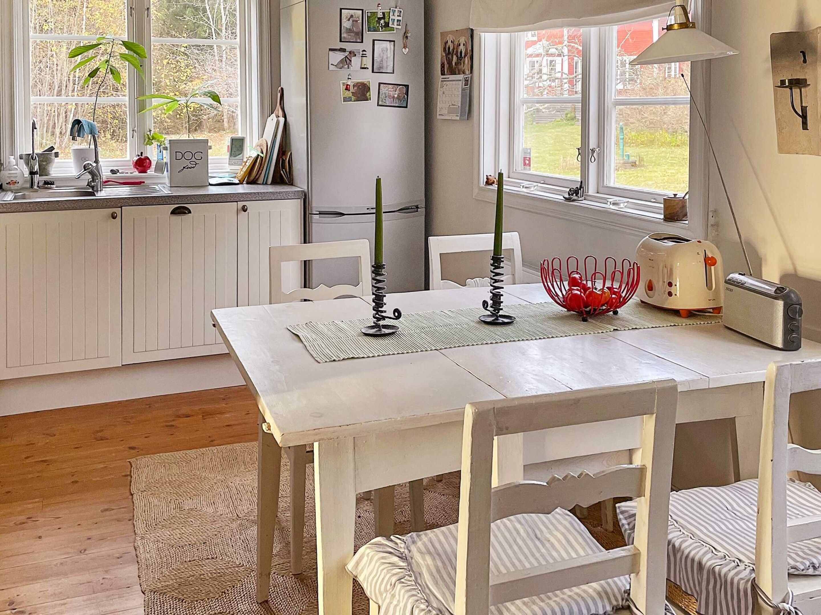 Una cocina con una mesa blanca, sillas, un tostador, una tetera y un ventanal con vistas al exterior.