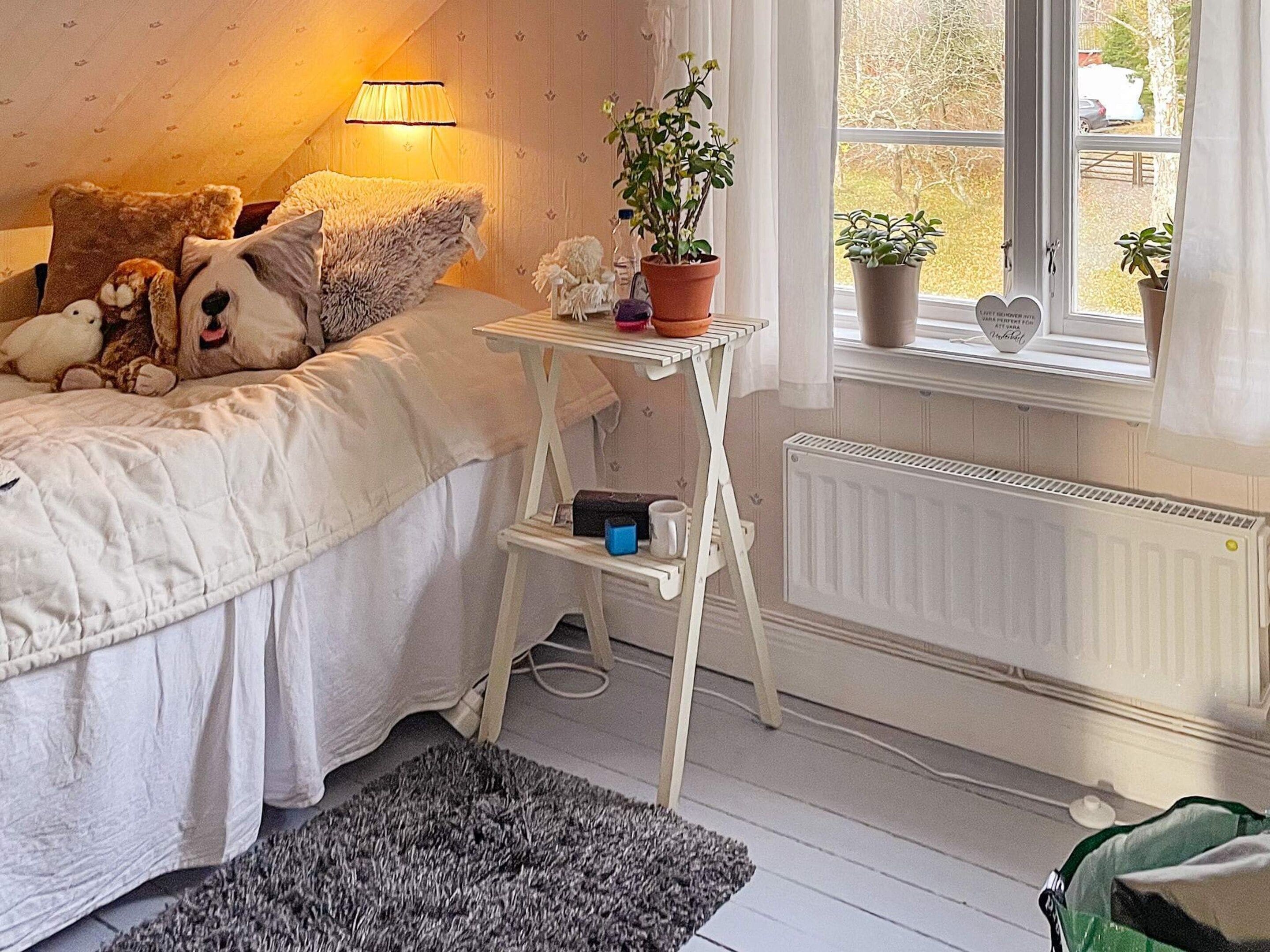 A bedroom with a bed, bedside table, lamp, potted plants, and a radiator.