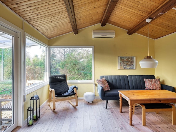 A room with a wooden ceiling, a sofa, a table, and a chair. There is a window providing a view of trees.