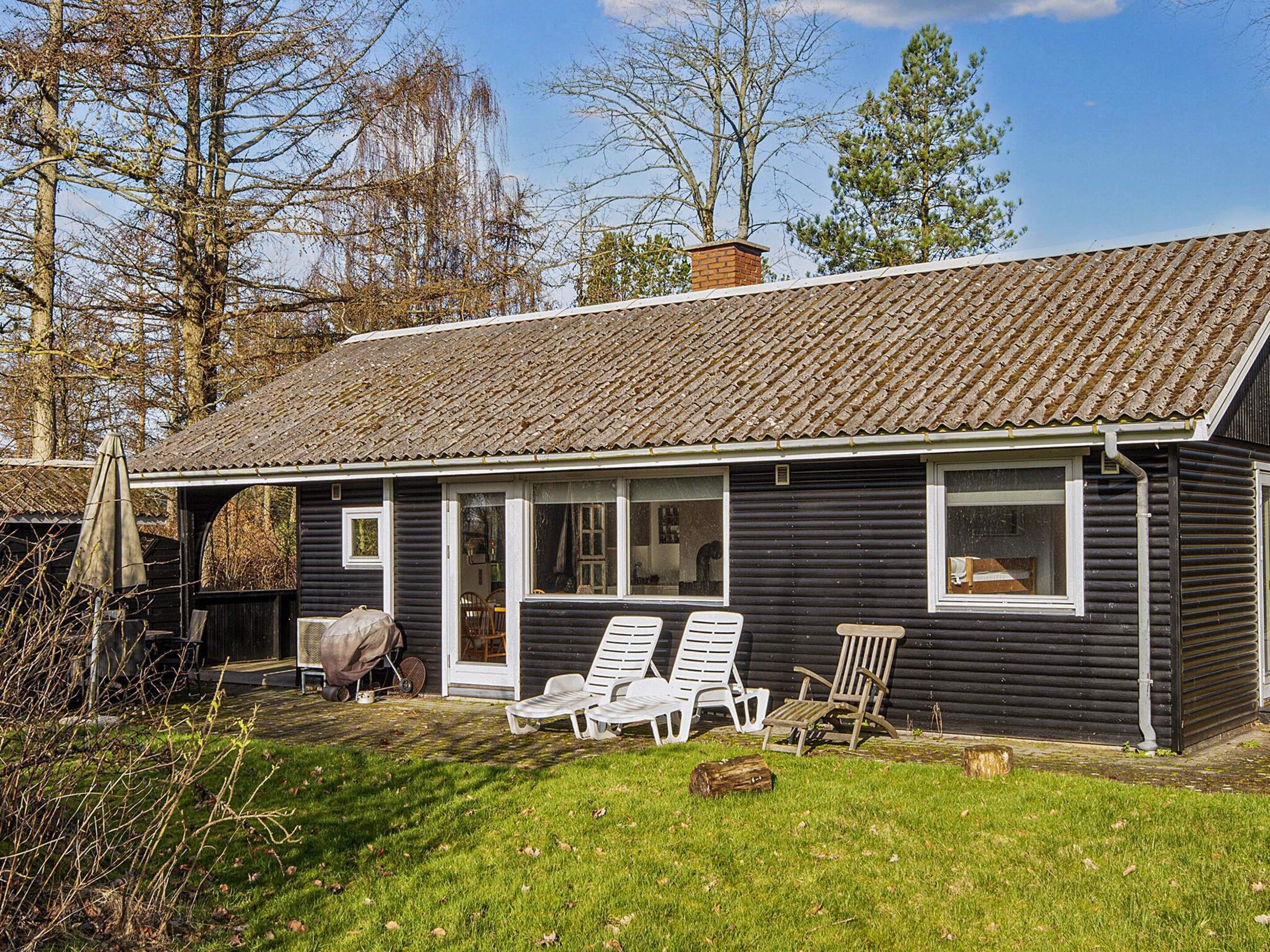 A small, dark wooden house with a tiled roof, a porch with white chairs, and a visible chimney.