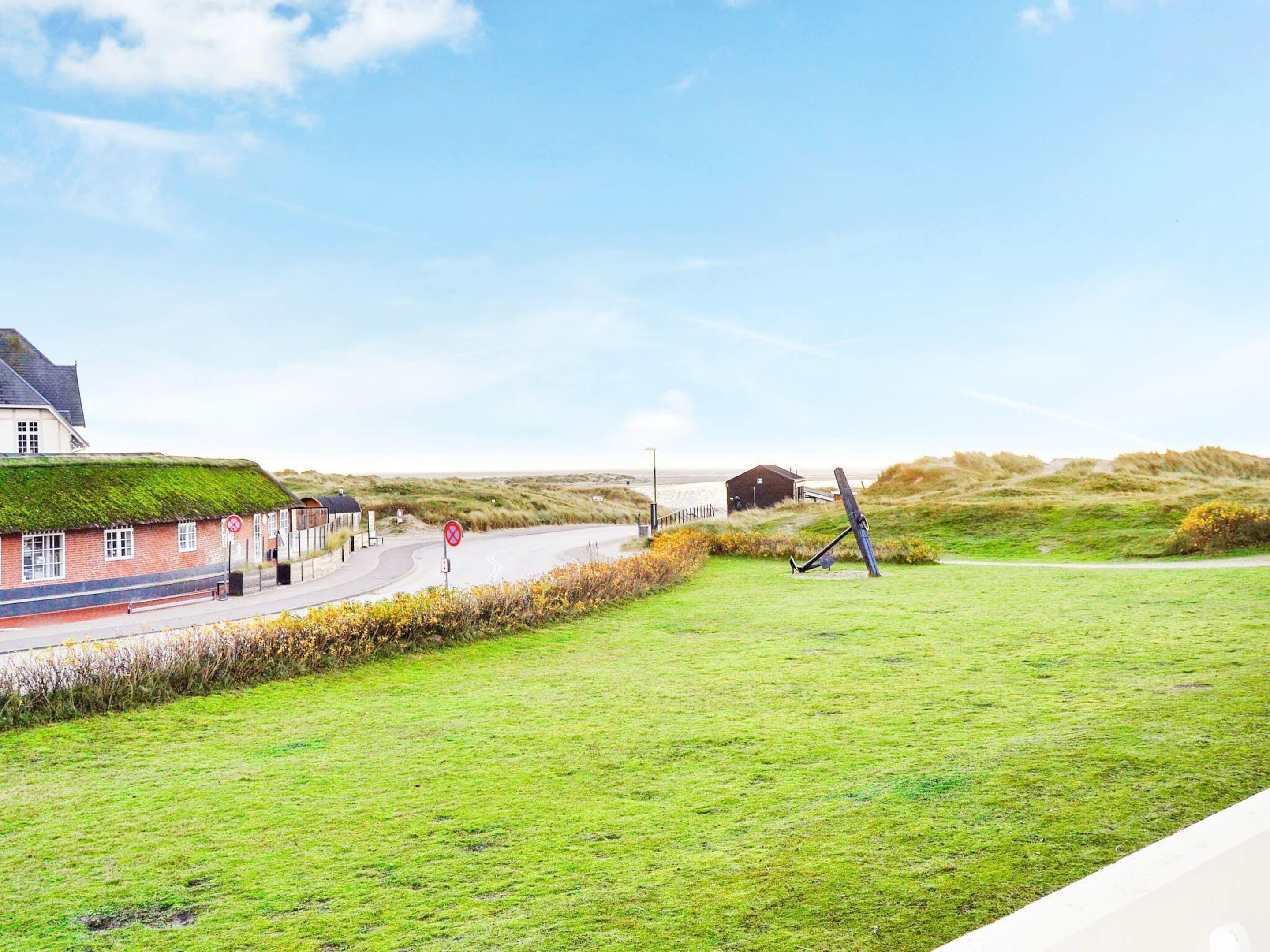 A coastal landscape with a building featuring a green roof, a winding road, and a grassy area with a sculpture.