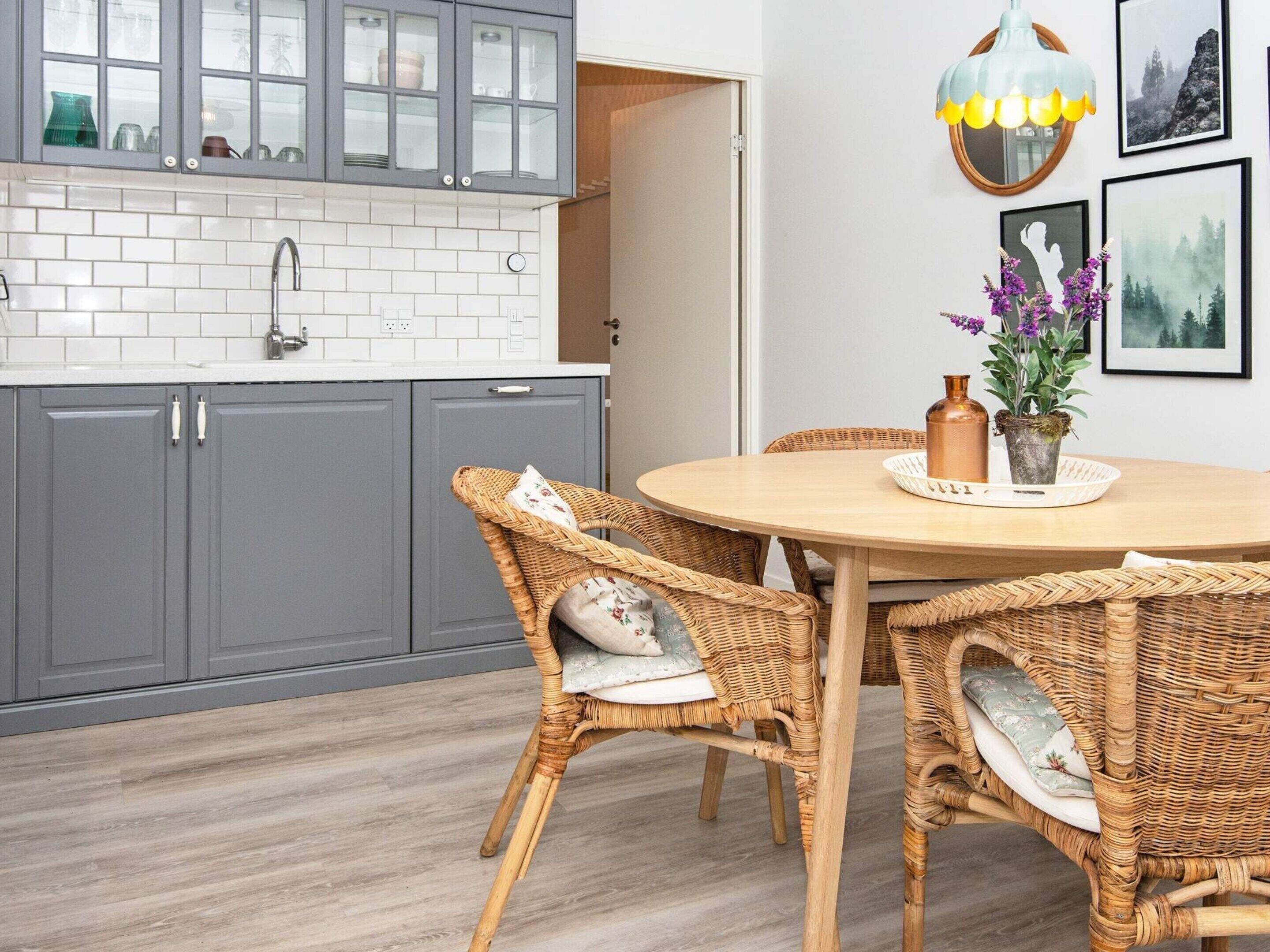 A kitchen with grey cabinets, a white tiled backsplash, and a round wooden table with wicker chairs.