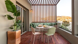 Ein Balkon mit Holzboden, einem Tisch mit Stühlen, einem Blumentopf, einer Couch mit Kissen und Blick auf eine hügelige Landschaft.