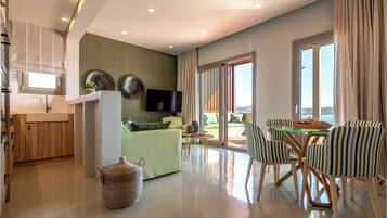 A modern living room with a sofa, dining table, and chairs, a kitchen area, and a large window with curtains.