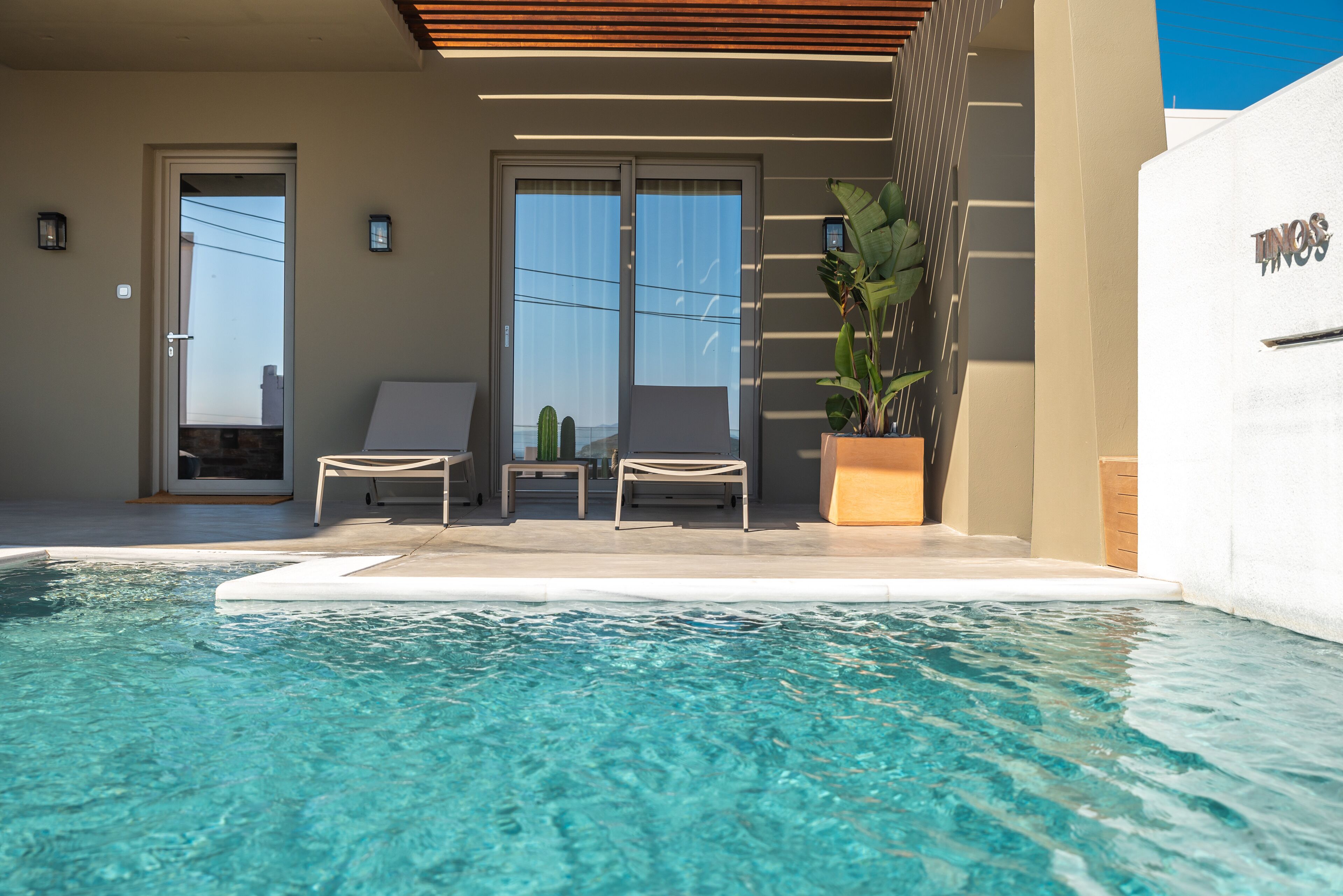 A modern pool area with clear water, two lounge chairs, a potted plant, and a sliding glass door.