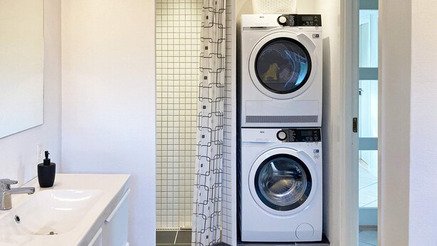 A compact laundry room with two stacked front-loading washing machines, a white sink, and a shower area with a patterned curtain.