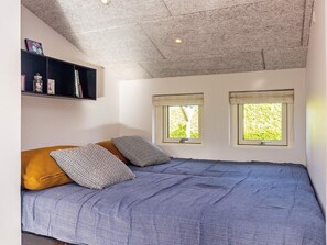 Un dormitorio con una cama, dos ventanas, un techo con textura y una estantería con libros y objetos decorativos.