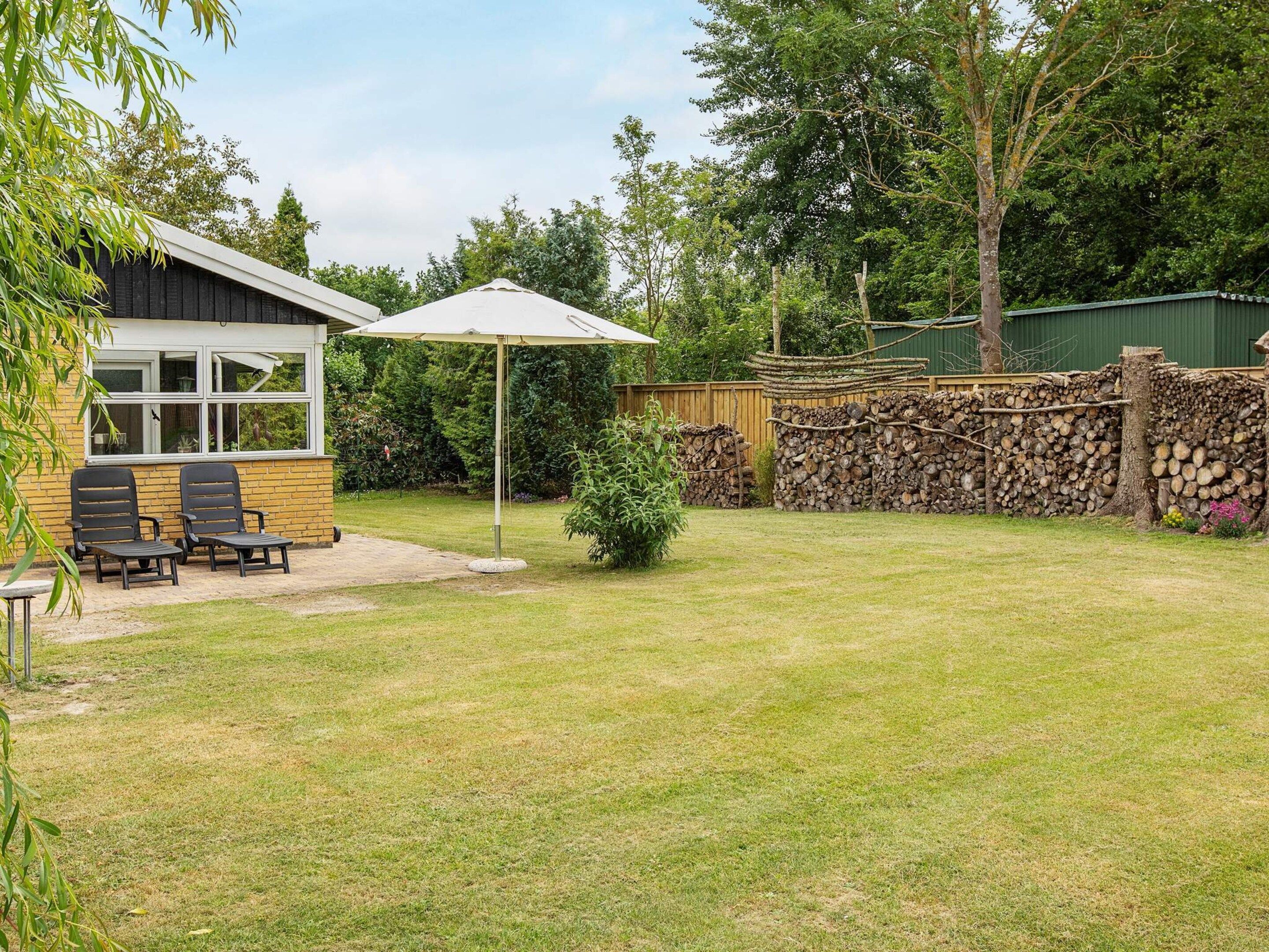 Un jardin avec une terrasse, deux chaises, une table, un parasol et une pile de bois de chauffage.