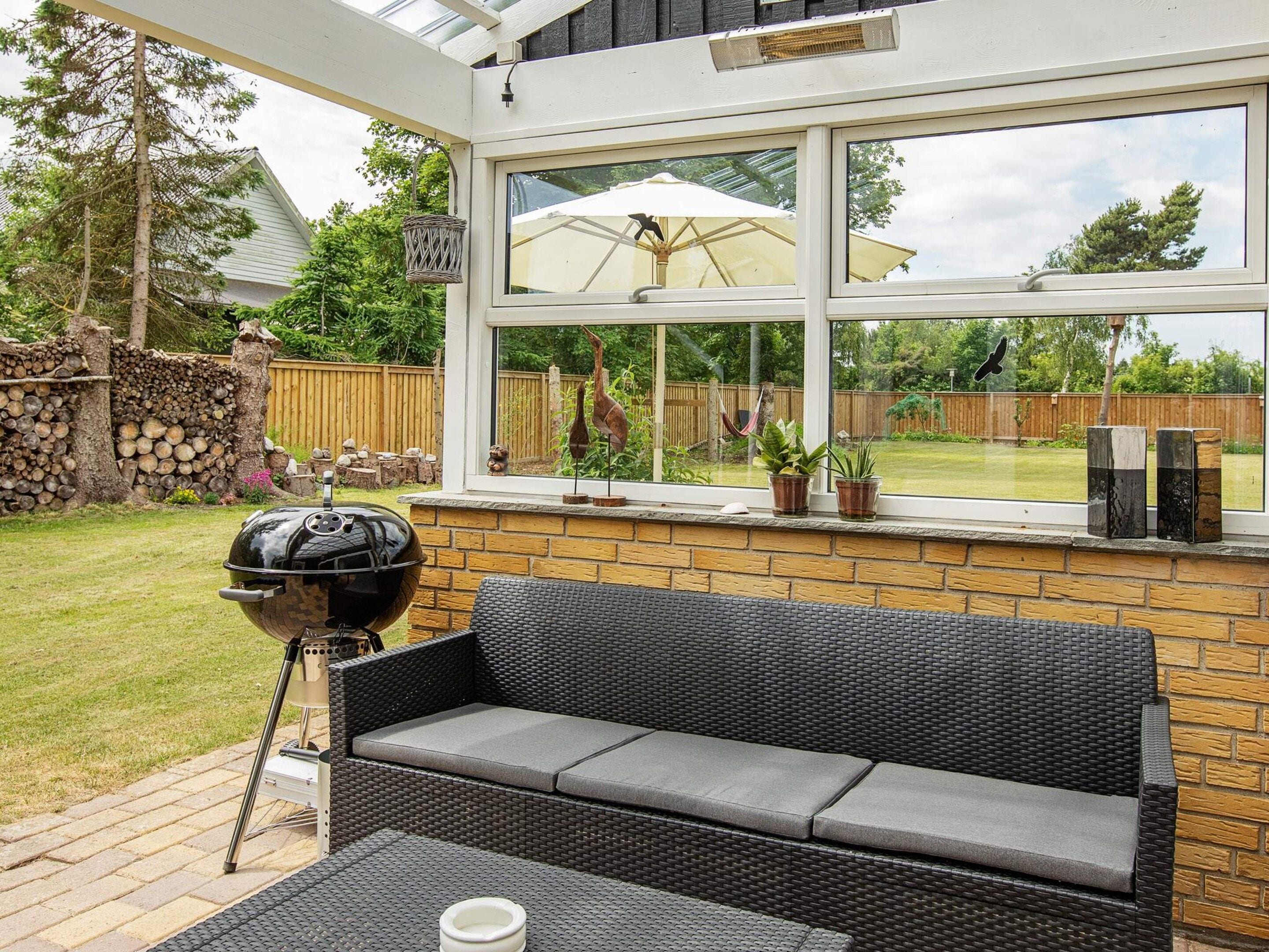 A patio with a black wicker sofa, a barbecue grill, and a view of a backyard through a large window.