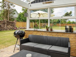 A patio with a black wicker sofa, a barbecue grill, and a view of a backyard through a large window. - 5 Person Holiday Home in Vaeggerlose (Vaeggerlose)