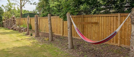 A hammock tied between trees in a backyard with a wooden fence and a stone wall.