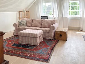 A cozy living room with a striped sofa, ottoman, and a red patterned rug. There's a wooden box labeled 'ACCOMPANIED BAGGAGE' and a lamp on a wooden shelf.