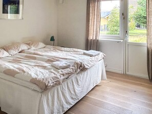 A bed with a white and beige bedspread, a folded towel, and a green floor lamp in a room with wooden flooring and a window with curtains.