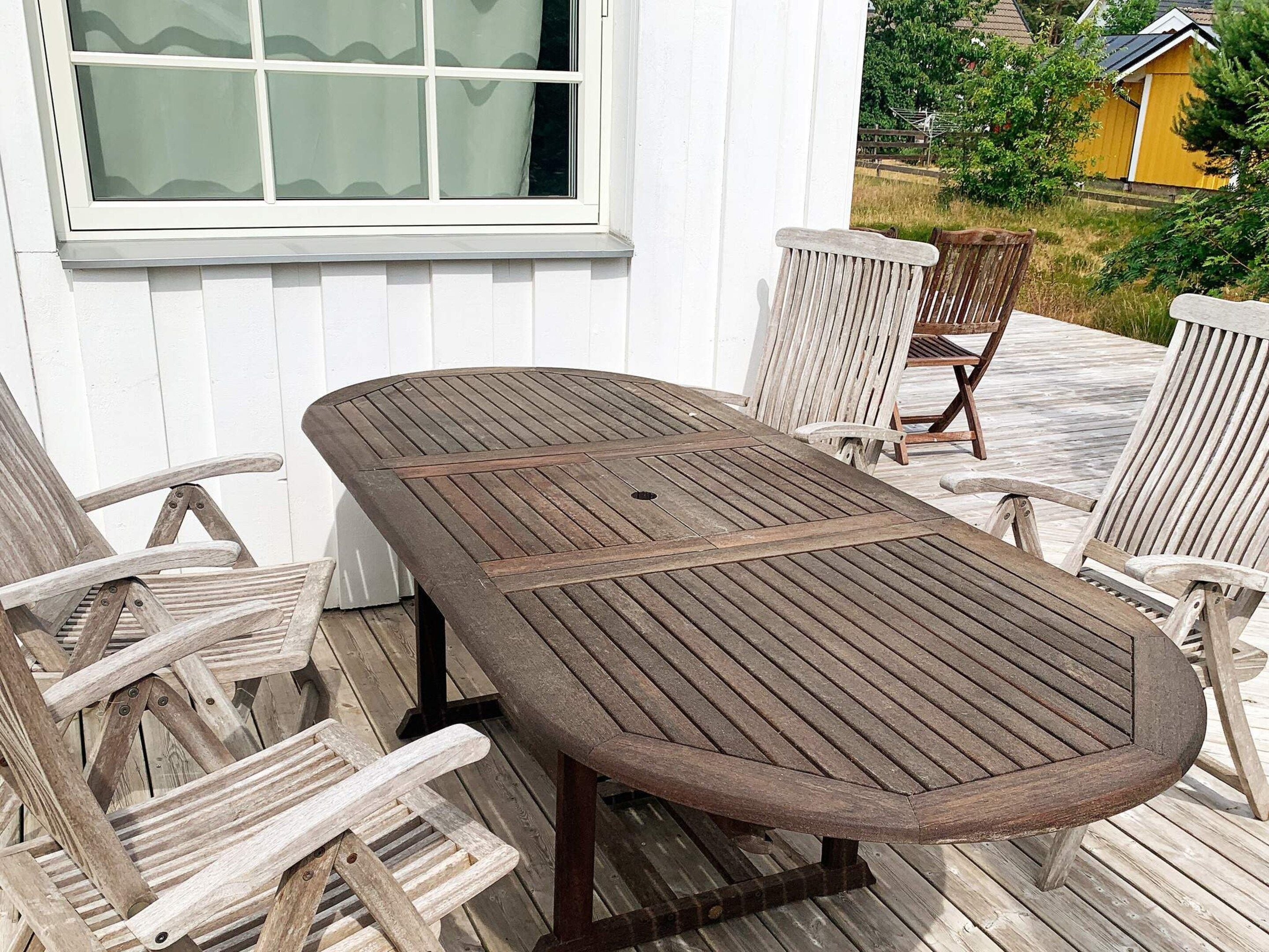 A wooden deck with a table and chairs, a window, and a view of a yellow building and greenery.