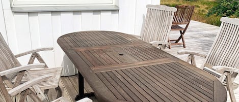 A wooden deck with a table and chairs, a window, and a view of a yellow building and greenery.