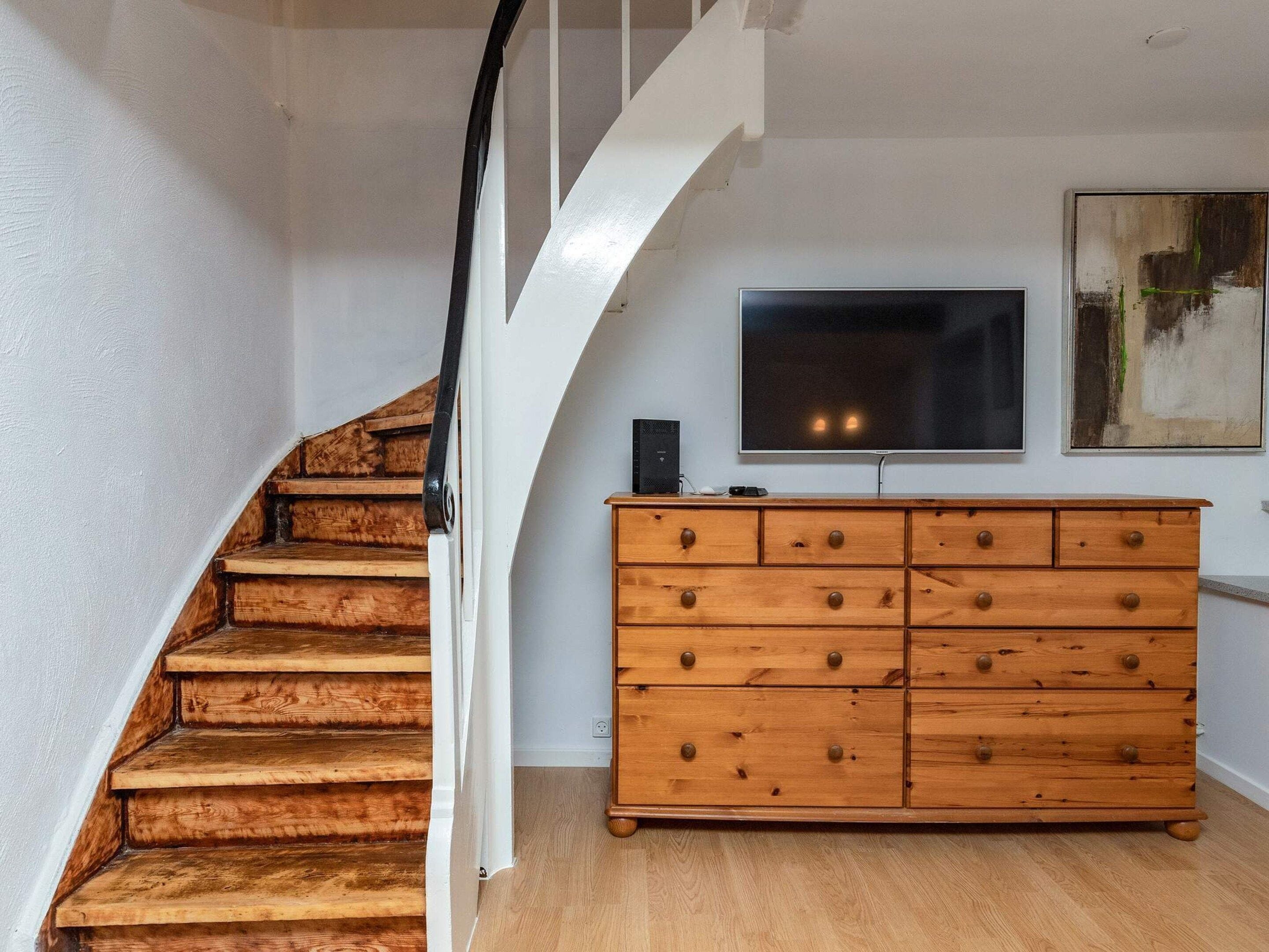 A staircase with wooden steps and a white wall, a wooden dresser with drawers, and a flat-screen TV mounted on the wall.