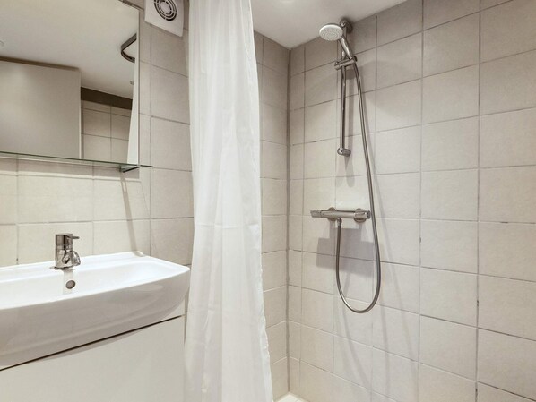 A bathroom with a white sink, a shower with a handheld showerhead, and a glass shelf above the sink.