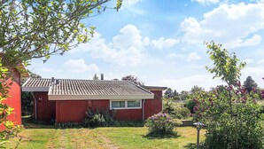 Une maison rouge avec un toit en tuiles, un petit jardin avec un chemin menant à l’entrée et un ciel bleu dégagé.