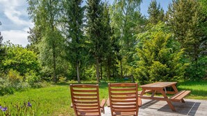 Een houten terras met twee stoelen en een picknicktafel, omgeven door weelderige begroeiing en bomen.