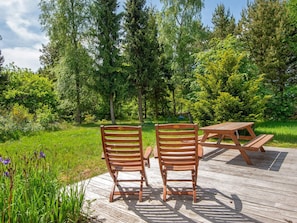 Une terrasse en bois avec deux chaises et une table de pique-nique, le tout entouré d’une végétation luxuriante et d’arbres.