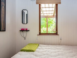 Un dormitorio con cama, ventana con vista a follaje, espejo y estante con tazas.