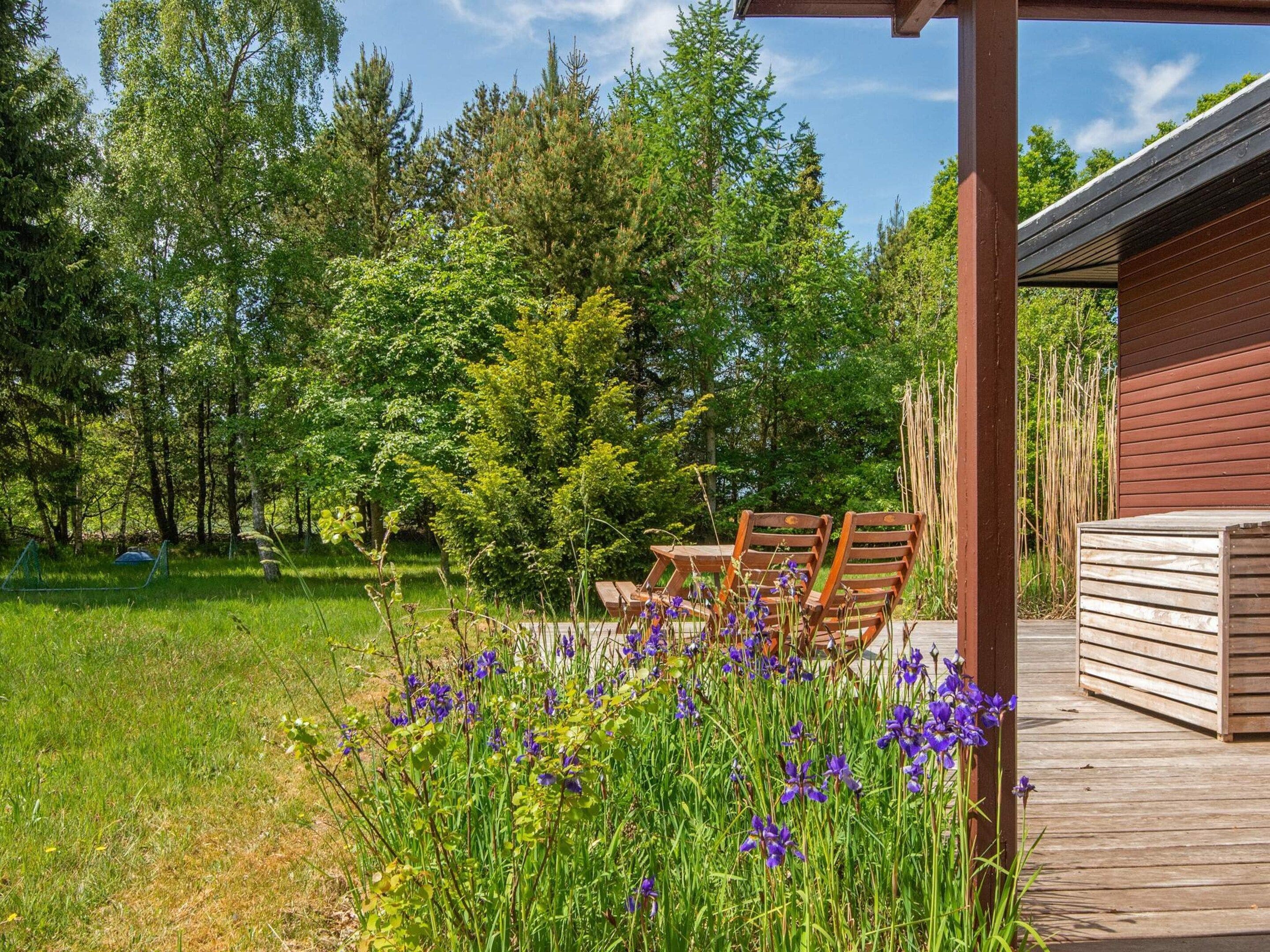 A wooden deck with two chairs, surrounded by a lush garden with purple flowers and tall trees.