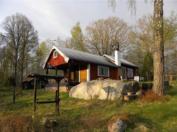 A rustic cabin with a sloped roof and a chimney, surrounded by trees and a large rock.