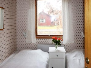 Quarto com uma cama, mesa de cabeceira com um vaso de flores e janela com vista para uma casa.