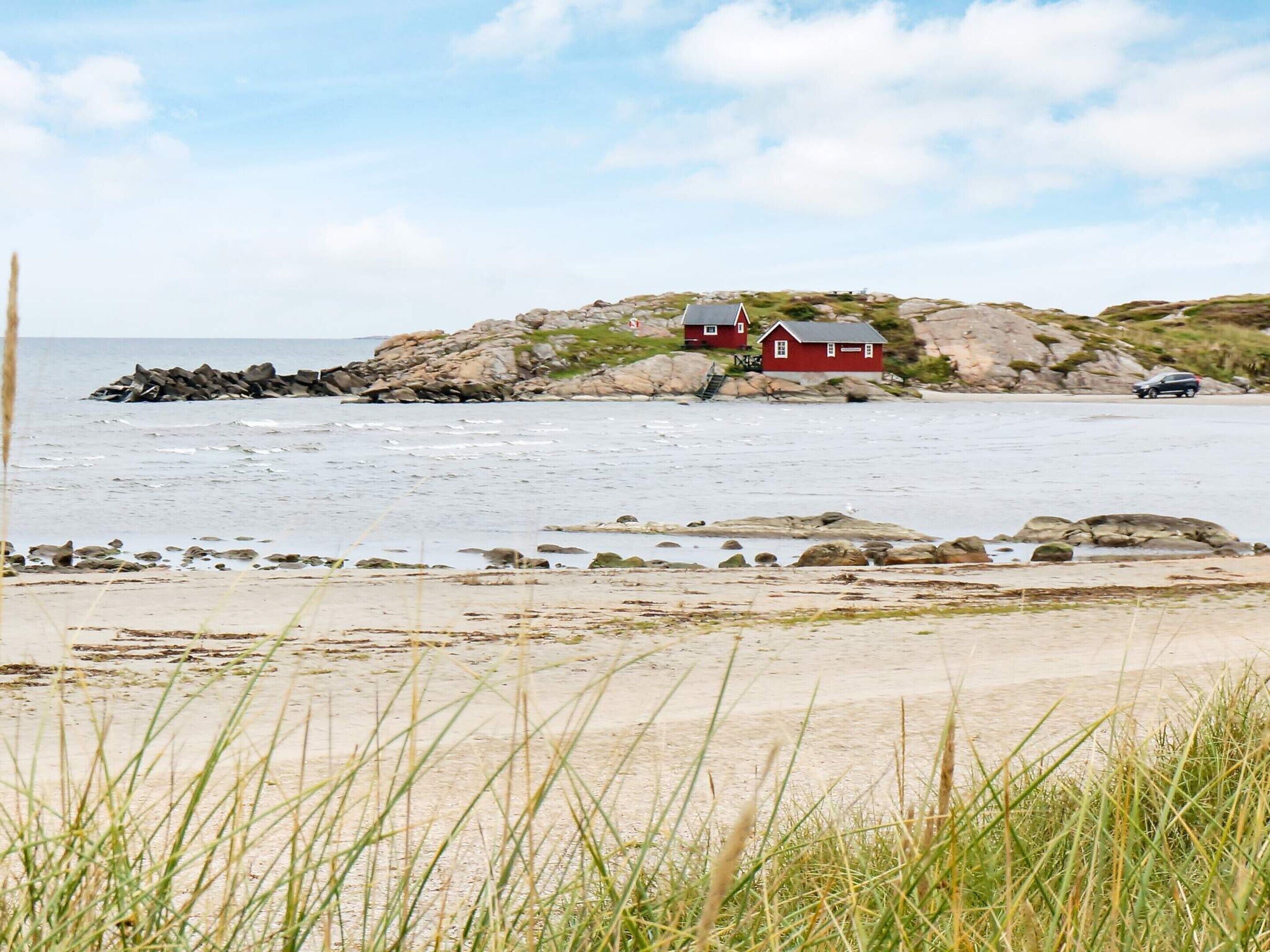 Uma paisagem costeira com uma casa vermelha em um afloramento rochoso, uma praia de areia e um mar calmo.