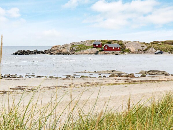 A coastal scene with a red house on a rocky outcrop, a sandy beach, and a calm sea.