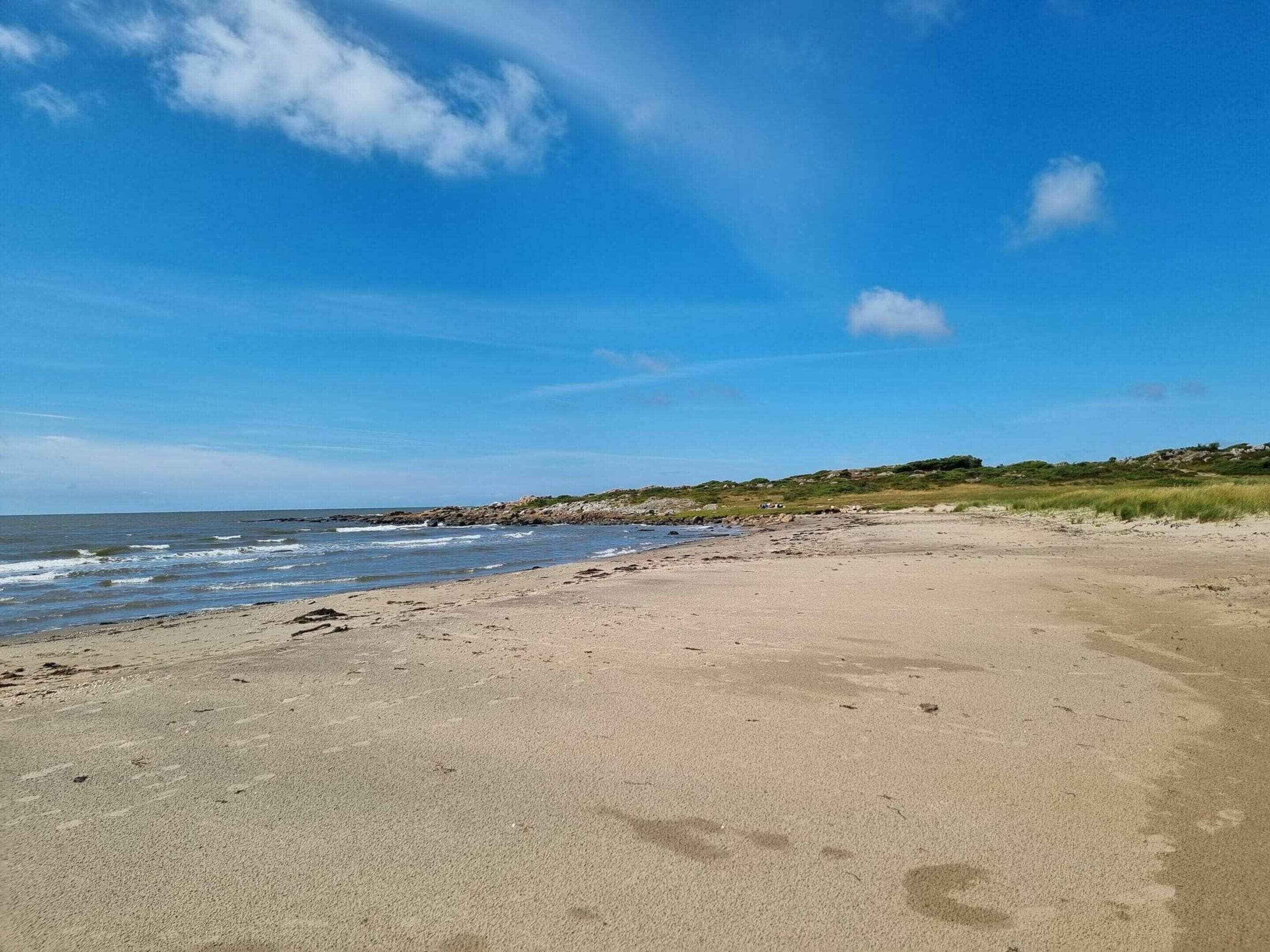 Une plage avec un ciel dégagé, un rivage sablonneux et des affleurements rocheux visibles au loin.